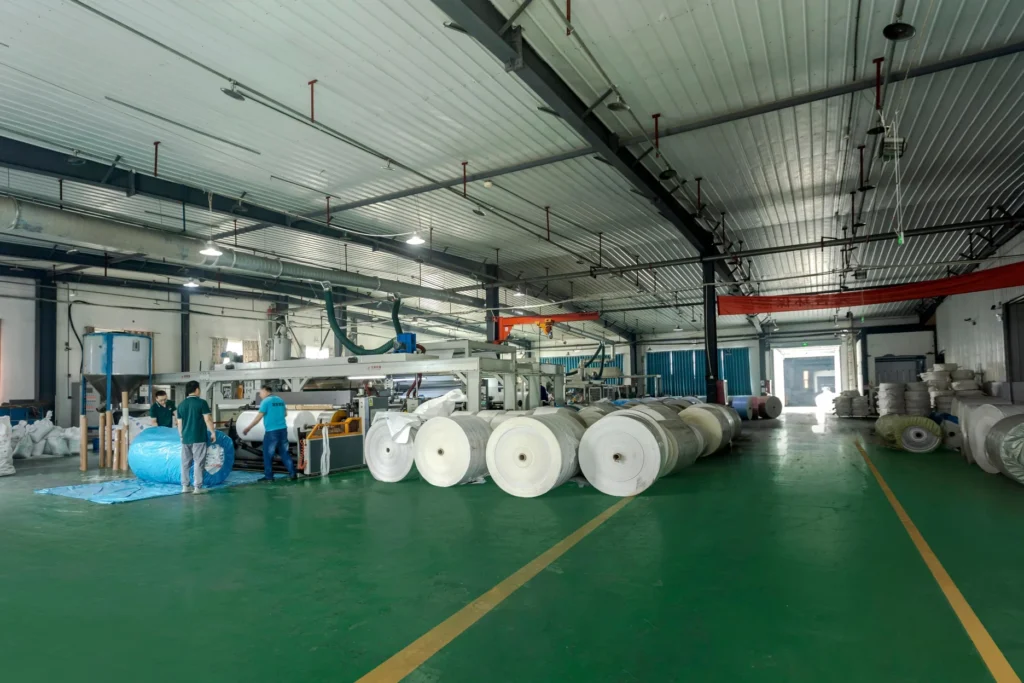 Interior view of an FIBC bulk bag manufacturing factory with woven fabric rolls and workers operating machinery.
