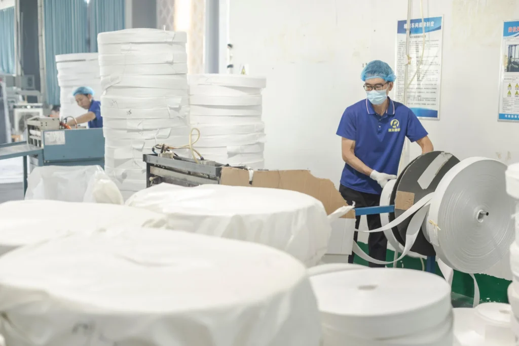 Workers in an FIBC bulk bag manufacturing factory handling woven fabric rolls.